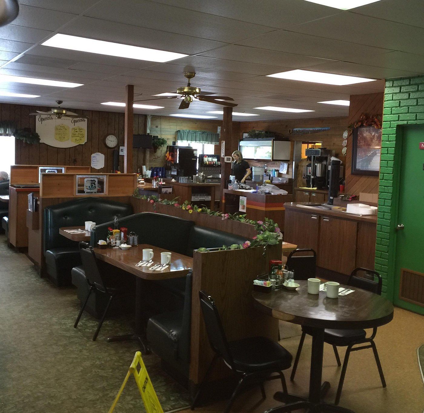 Dining room with dark green booths, round tables, wood-paneled counter, ceiling fans and a green brick wall